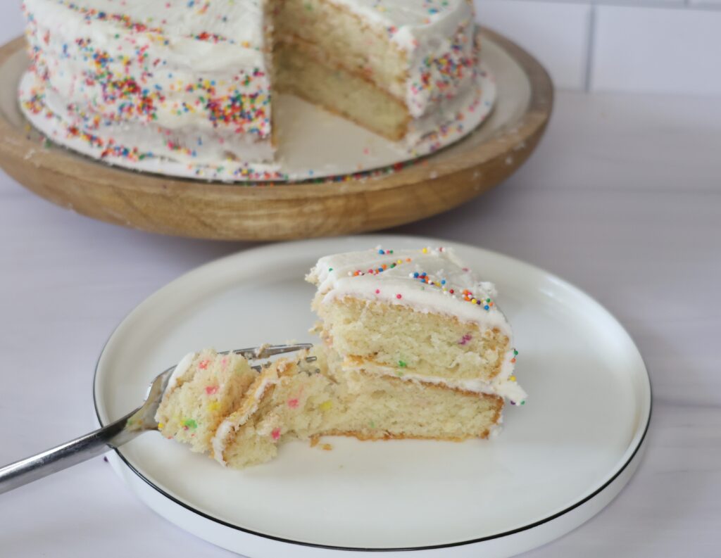 A white plate with a decrative slice of vanilla birthday cake sitting next to a larger cut birthday cake.