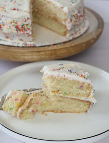 A white plate with a decrative slice of vanilla birthday cake sitting next to a larger cut birthday cake.