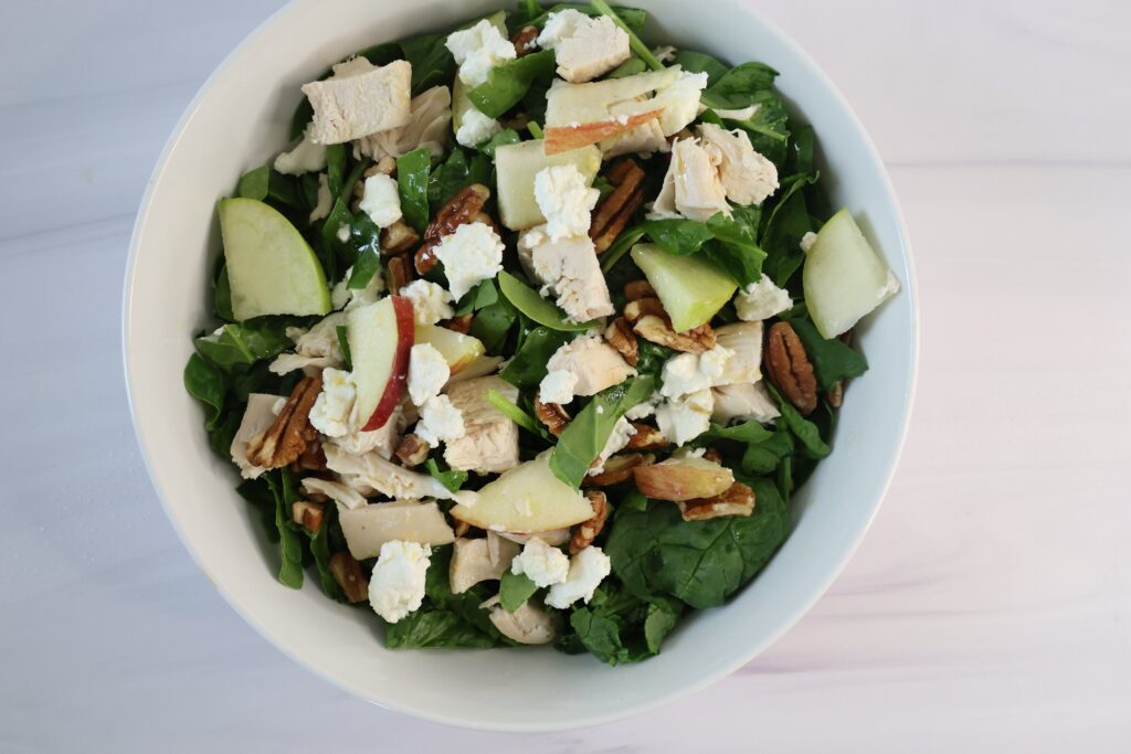 A green salad with chicken,apples and white goat cheese in a white bowl.