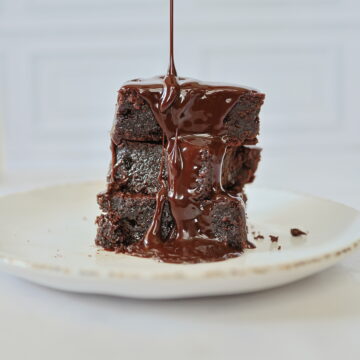 three brownies stacked on a white plate with syrup dripping over them.