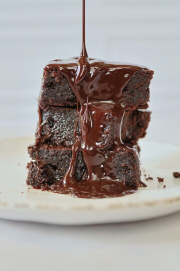 three brownies stacked on a white plate with syrup dripping over them.