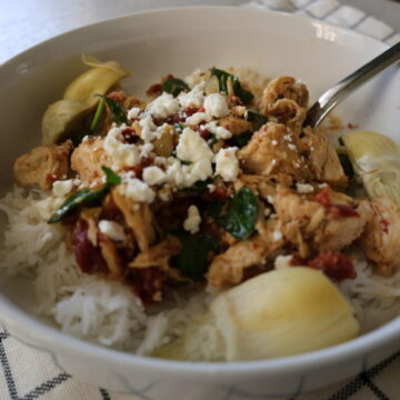 Juicy chicken and artichokes topped with sundried tomatoes, spinach and feta in a white bowl with a metal spoon.
