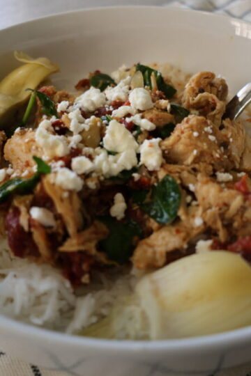 Juicy chicken and artichokes topped with sundried tomatoes, spinach and feta in a white bowl with a metal spoon.