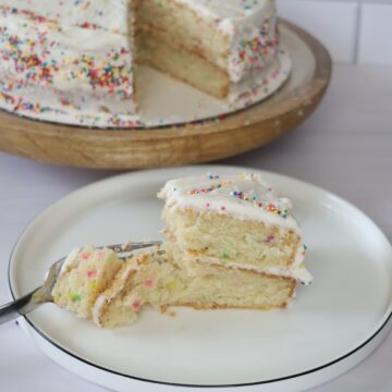 A slice of gluten-free birthday cake sitting on a small white plate, placed in front of a full cake with one slice missing.