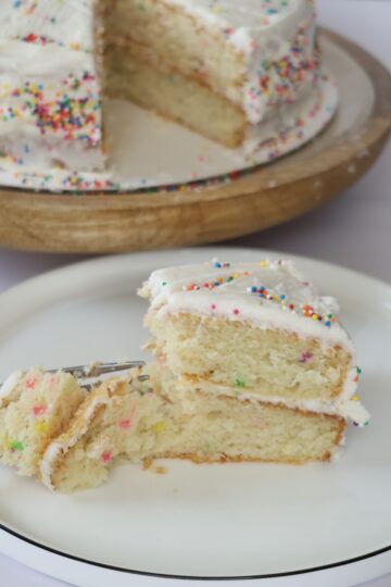 A slice of gluten-free birthday cake sitting on a small white plate, placed in front of a full cake with one slice missing.