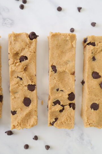 four homemade peanut butter protein bars with chocolate chips laying on a white surface with chocolate chips sprinkled around them.