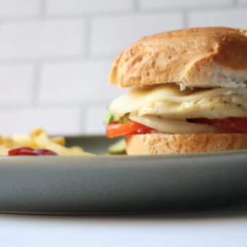 A blue/gray plate with an in focus grilled chicken sandwich with tomatoes, avocado and gluten-free bun and out of focus french fries.