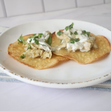 Two yellow corn tostadas with creamy chicken and cilantro on a white plate laying over a blue and white napkin.