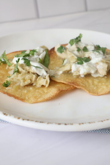 Two yellow corn tostadas with creamy chicken and cilantro on a white plate laying over a blue and white napkin.