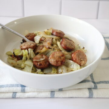 Sliced sausage with green cabbage and onions in white bowl with a silver spoon.