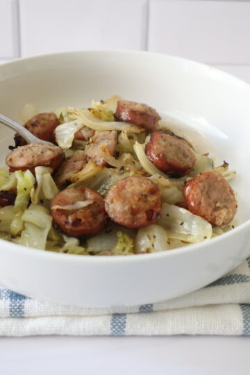 Sliced sausage with green cabbage and onions in white bowl with a silver spoon.