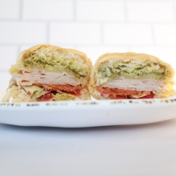 A white plate with a gluten-free sub sandwich cut in half. Sandwich contains green pesto, white turkey and red pepperonis.