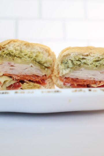 A white plate with a gluten-free sub sandwich cut in half. Sandwich contains green pesto, white turkey and red pepperonis.