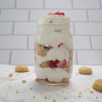 A clear mason jar with layers of white whipped cream, strawberries and shortbread cookies.