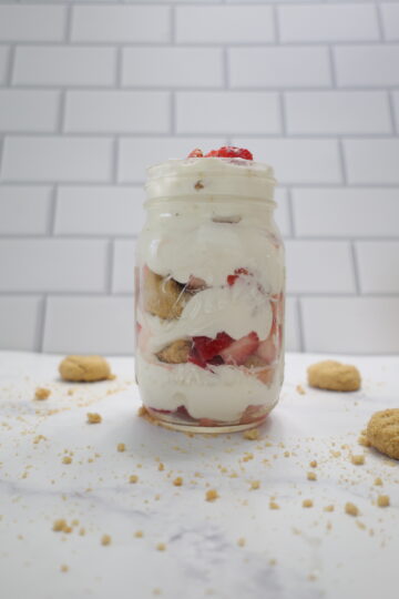 A clear mason jar with layers of white whipped cream, strawberries and shortbread cookies.