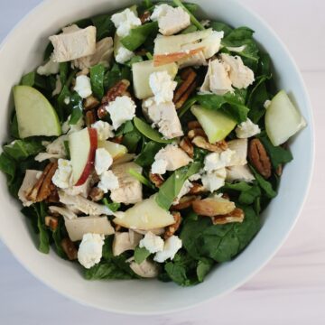 A medium white bowl containing bright green greens, red and green apples, white goat cheese, chicken and a small amount of pecans.