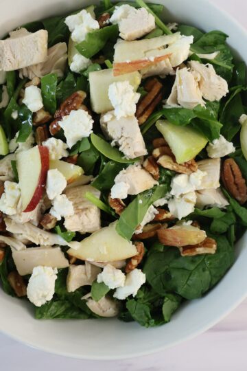 A medium white bowl containing bright green greens, red and green apples, white goat cheese, chicken and a small amount of pecans.