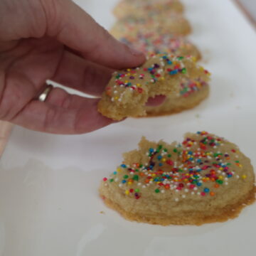 Almond flour cookies with rainbow sprinkles lining a white pan with one almond flour cookie broken in half.