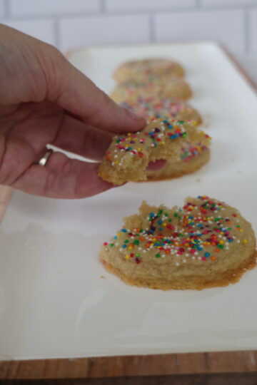 Almond flour cookies with rainbow sprinkles lining a white pan with one almond flour cookie broken in half.