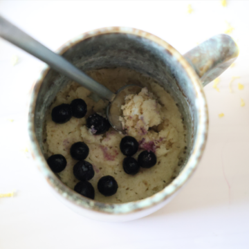 A lemon and almond flour bake sprinkled with blueberries filling half of a light blue mug.