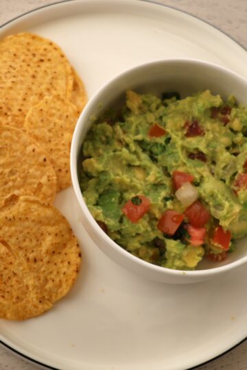 A white plate containing tortilla chips on the left and a small white bowl of light green guacamole with diced red tomatoes scattered throughout