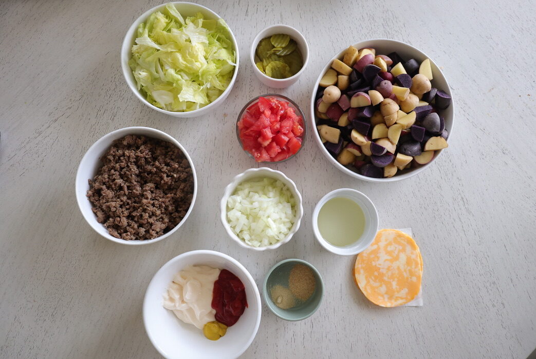 Lettuce, pickles, tomatoes, ground beef, onions, potatoes, oil, spices, ketchup, mayo and mustard on a white surface.