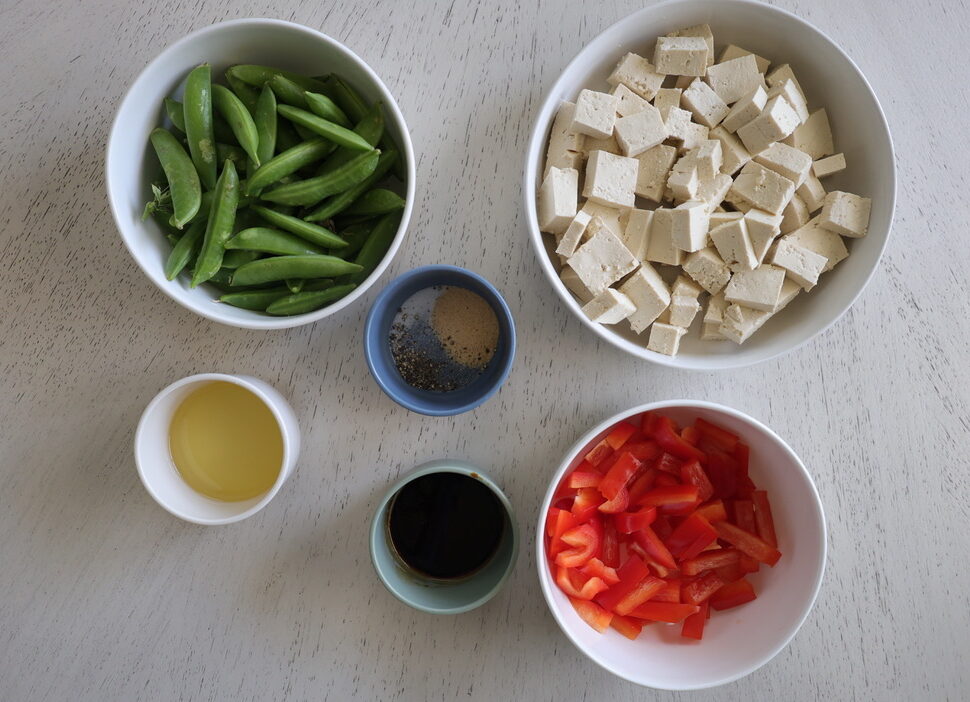 A white background with bowls of tofu, snap peas, red bell peppers, oil, soy sauce and spices.