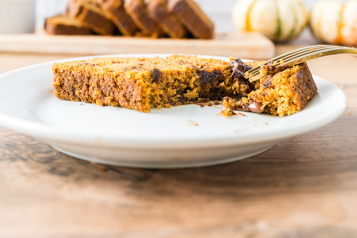 A slice of gluten-free almond flour pumpkin bread with chocolate chips on a white plate with one bite on a fork. Yellow and orange pumpkins in the background.