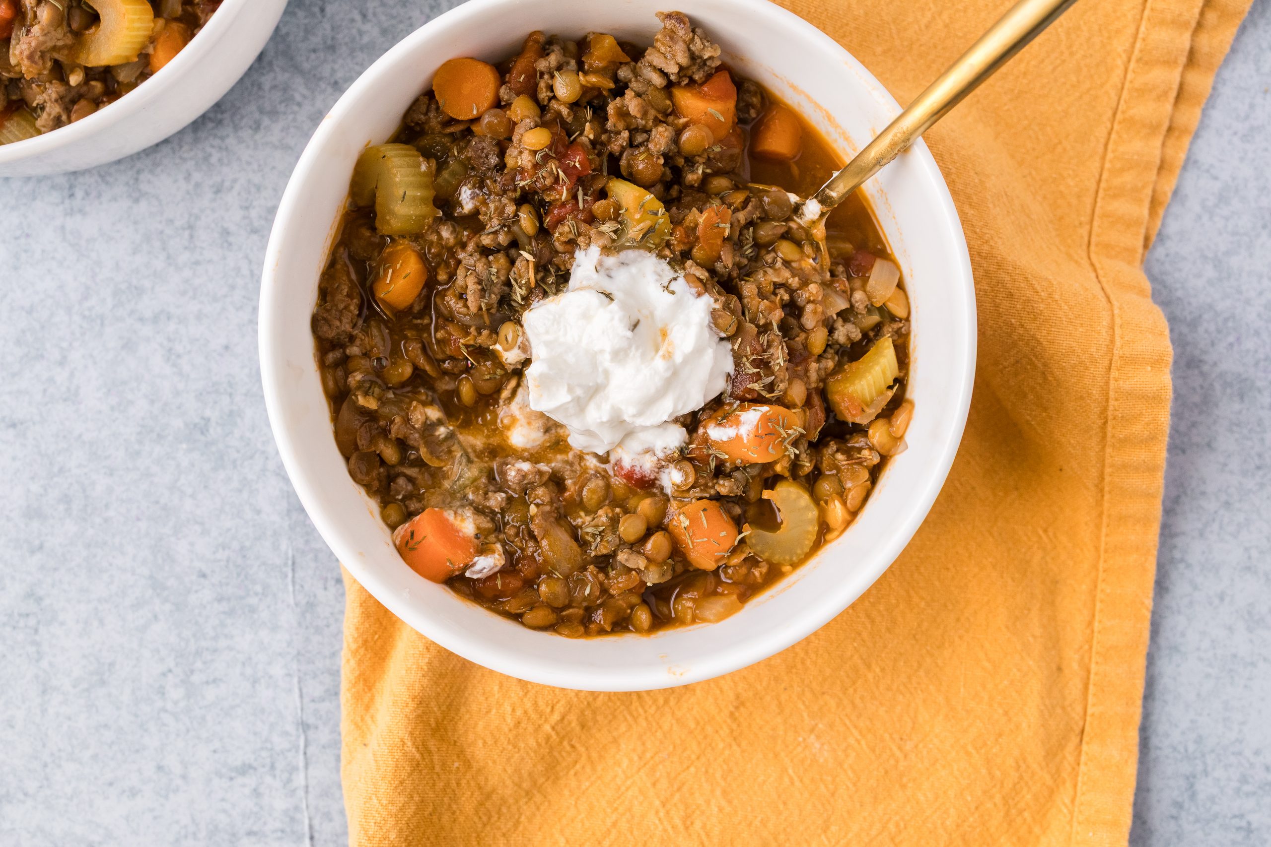 Beef, lentil and vegetable soup served in a white bowl with sour cream on top, a gold spoon and a yellow and blue background. 
