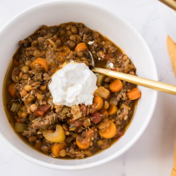Beef lentil and vegetable soup served in a white bowl with a gold spoon and topped with sour cream.