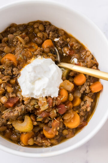 Beef lentil and vegetable soup served in a white bowl with a gold spoon and topped with sour cream.