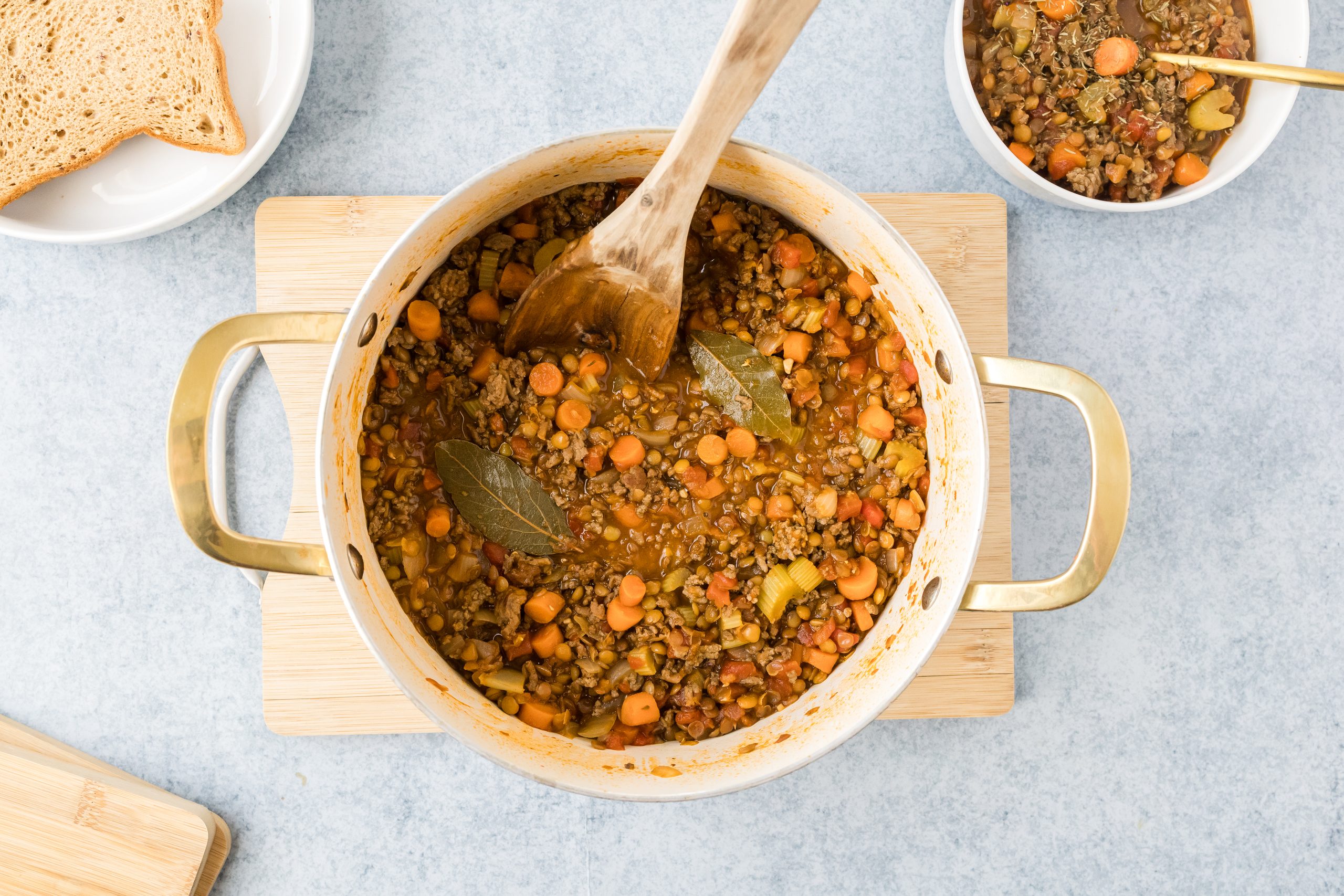 Beef lentil and vegetable soup served in a large white bowl with a wooden spoon over a wooden cutting board and blue background. 